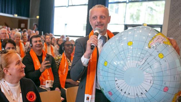 Mann mit einer großen Weltkugel unter dem Arm spricht vor einer Versammlung